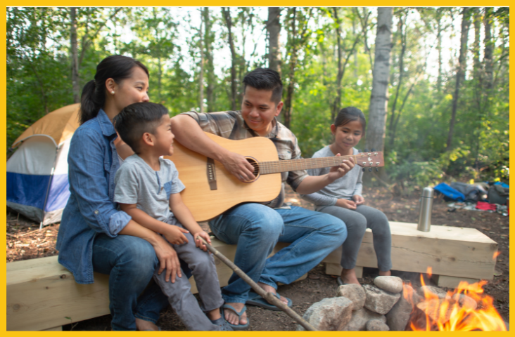A family out in nature enjoying their family tradition of singing and playing the guitar around the campfire.