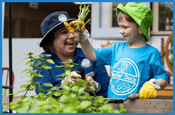 Early childhood educator in Mother Duck Childcare uniform in the garden with a young boy holding up a plant